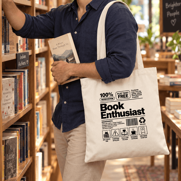 A man in a bookstore stands by a bookshelf, holding a book and wearing a white canvas tote bag printed with the words 'Book Enthusiast' and small icons. The tote bag has a black printed design and is shown in a warm, real-world bookstore environment.